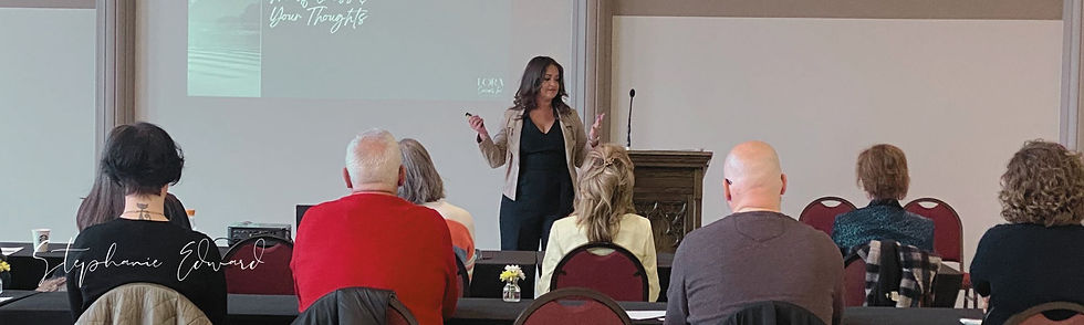 Stephanie Edward is a white woman with brown hair and brown eyes. She is wearing a dark jumpsuit and beige jacket. She is speaking in front of a screen to a group of seated people,