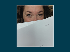 Stephanie Edward is a white woman. She is holding up her manuscript and peeking out from behind it.
