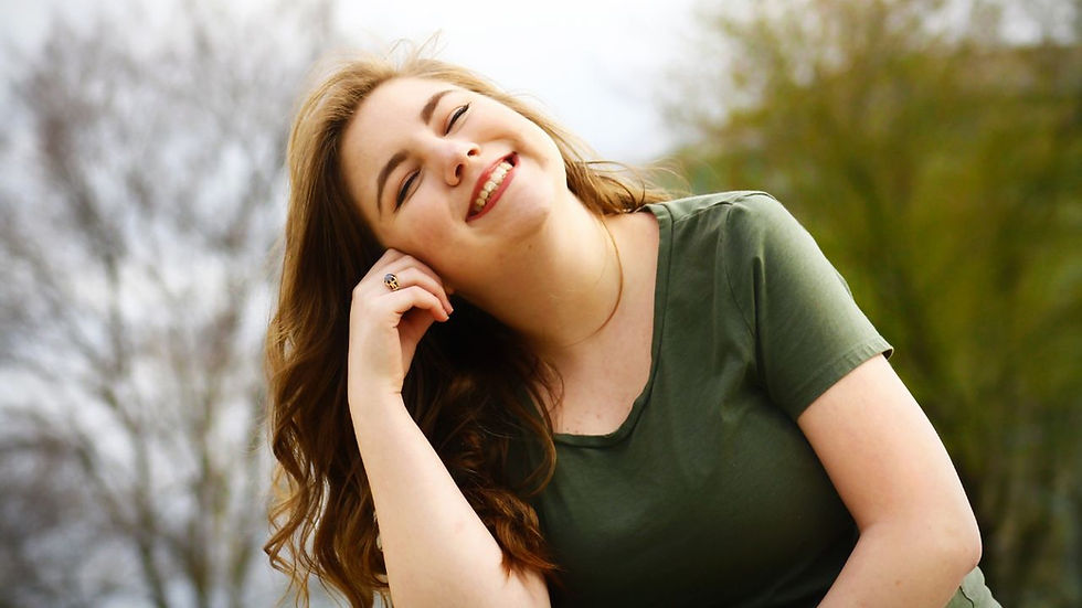 Smiling woman in a green shirt leans on hand, outdoors with blurred trees in background. Bright and cheerful mood.