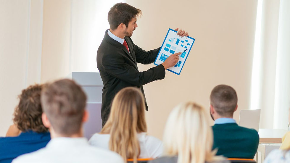 Man in a suit presenting a blue chart to an audience in a bright room. The audience is seated and attentive, creating a professional mood.