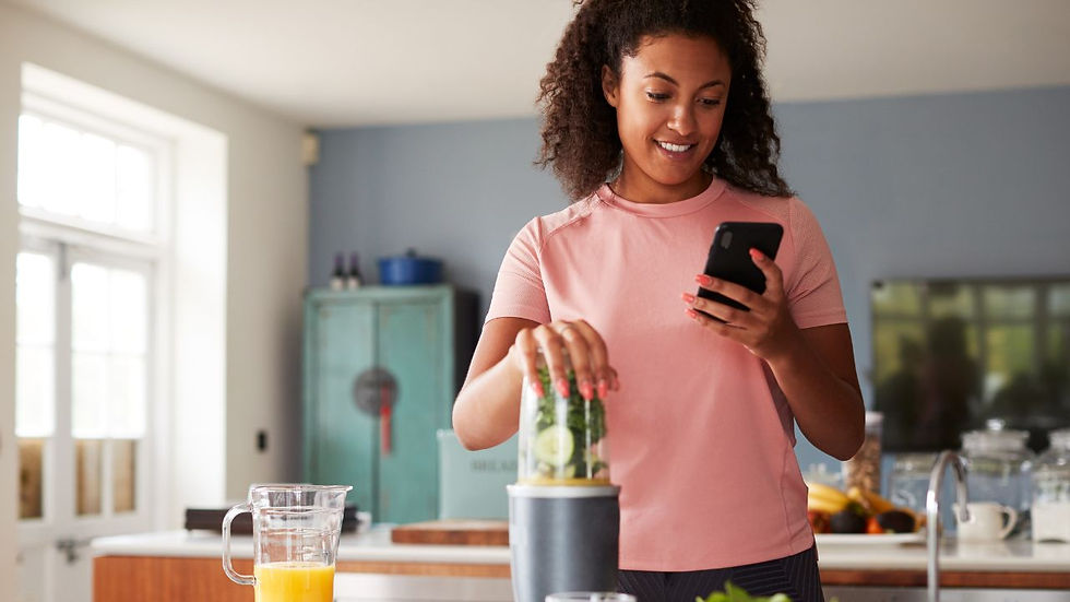 Woman in a pink shirt uses a blender with cucumber inside while checking her phone in a bright kitchen. Glass of orange juice nearby. Smiling.