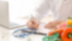 Doctor in white coat writing on clipboard, laptop nearby. Stethoscope, fresh fruits, and measuring tape on table. Bright, office setting.