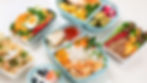 Various meal prep containers with colorful dishes including salmon, rice, veggies, pasta, and fruit. A vibrant, healthy food display.