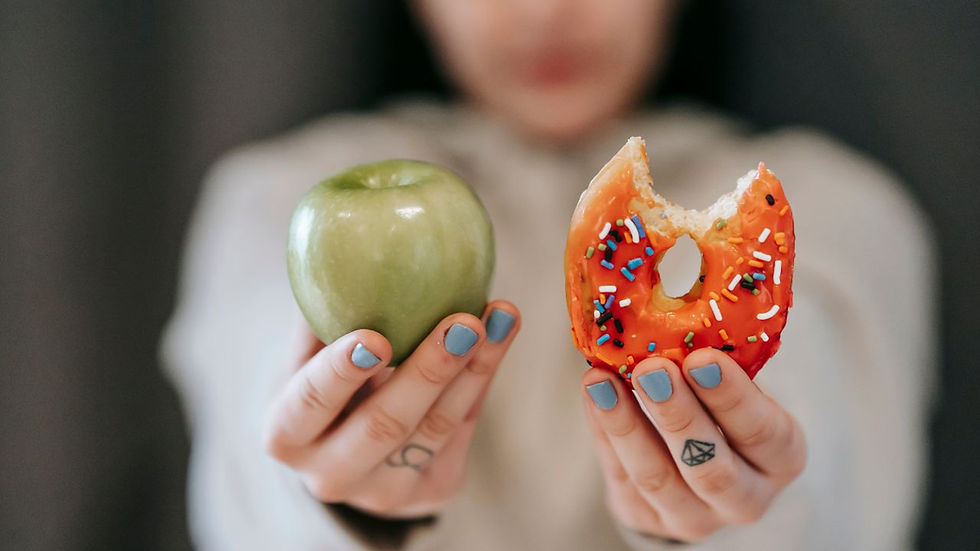 A person holds a green apple and a bitten donut with sprinkles, showing blue nail polish. The background is blurred.