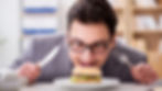 Man in glasses eagerly looks at a burger with a knife and fork in hand, in an office setting. Bright, focused expression.
