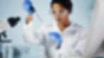 Scientist in goggles and white lab coat using a pipette in a lab. A microscope and test tubes are nearby. Focused and precise.