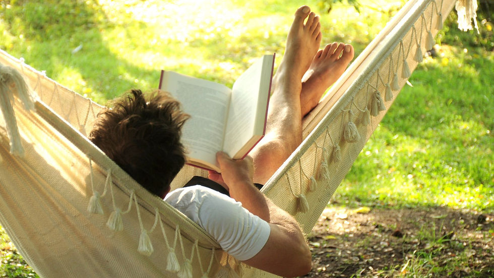 Person relaxing in a hammock, reading a book in a sunny garden. The hammock is beige with tassels. Peaceful and serene mood.