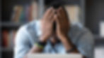 Person with head in hands, wearing a blue patterned shirt and bracelets, appears stressed at a desk. Blurred bookshelf in the background.