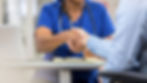 A person in blue scrubs shakes hands with someone in a blue shirt across a desk with a laptop and clipboard. Professional setting.