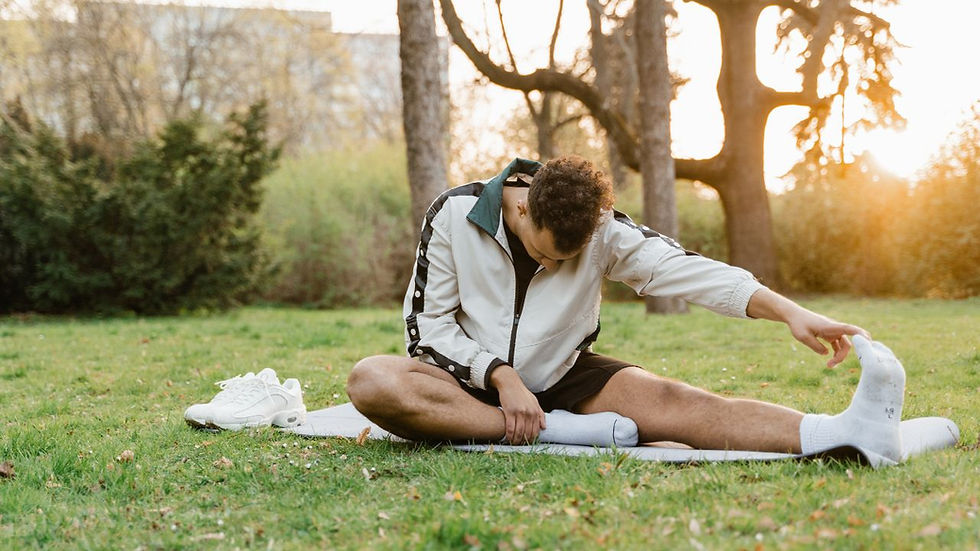 Person stretching on grass in park, reaching for feet. Wearing a white jacket and shorts. Trees and sunset in background. Relaxed mood.