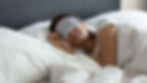 Woman sleeping peacefully in bed, wearing a striped sleep mask. White bedding, textured dark headboard in background, serene mood.