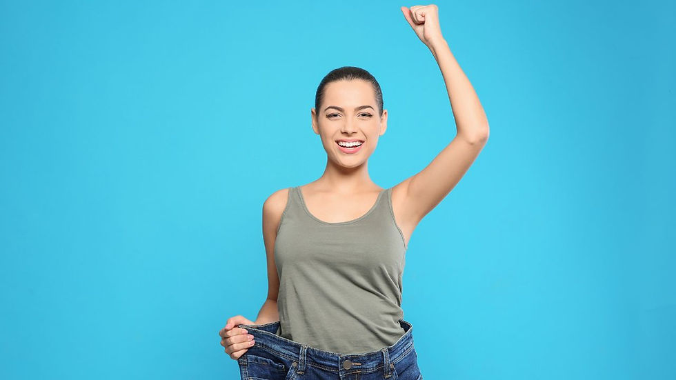 Woman in a gray tank top smiles, holding oversized jeans and raising a fist in triumph against a bright blue background.