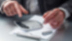 Hands in a suit operate a calculator on a clipboard with paper. A pen rests nearby. The setting is a metallic desk, suggesting a business mood.