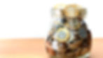 A glass jar filled with assorted coins, predominantly one and two-pound coins, is placed on a wooden surface. The coins are stacked and overlapping, creating a reflective sheen. The background is plain white, emphasizing the jar.