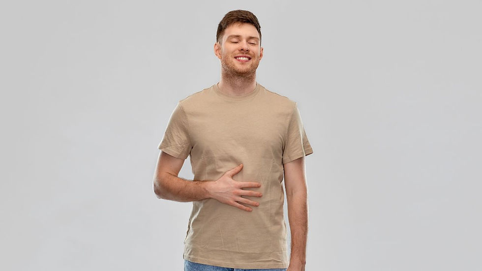 Man in a beige shirt smiling with eyes closed, hand on stomach, against a plain gray background. Casual, content mood.