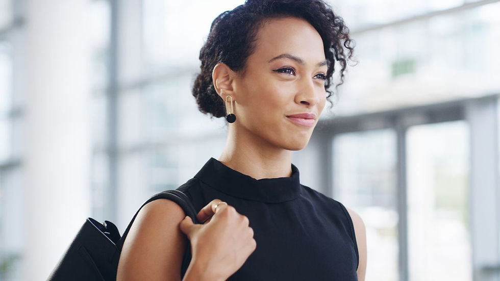 Woman in black dress smiles confidently, holding a bag over shoulder, in a bright, modern office setting. Background is softly blurred.