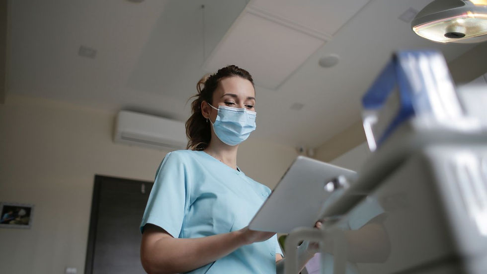 Nurse in blue scrubs and mask, reading a clipboard in a medical setting. Calm mood, neutral-colored room with ceiling light.