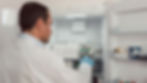 Man in white lab coat and blue gloves examines vials inside a lab fridge. Shelves hold boxes and containers. Bright, clinical setting.