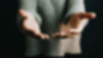 Pair of open hands reaching out, palms up, against a dark background. Person wears a gray sweater, conveying a gesture of offering.