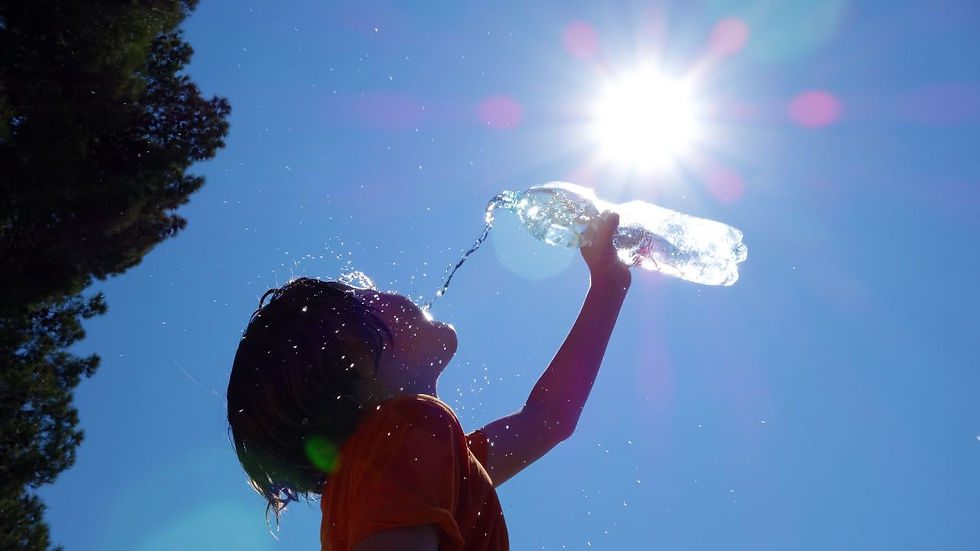 Child in orange shirt drinks from a water bottle under bright sun in a blue sky. Water droplets sparkle, creating a joyful summer vibe.