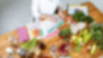 Person planning a meal with charts, surrounded by fruits, vegetables, and kitchen tools on a wooden table. Vibrant, organized setting.