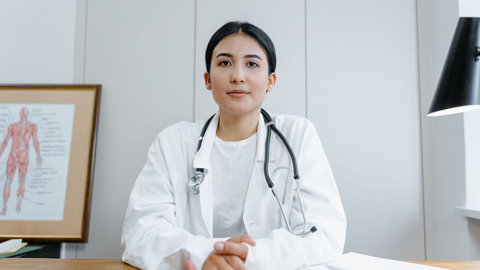 Doctor in white coat with stethoscope sits at desk, neutral expression. Anatomical poster on the wall, soft lighting from lamp.