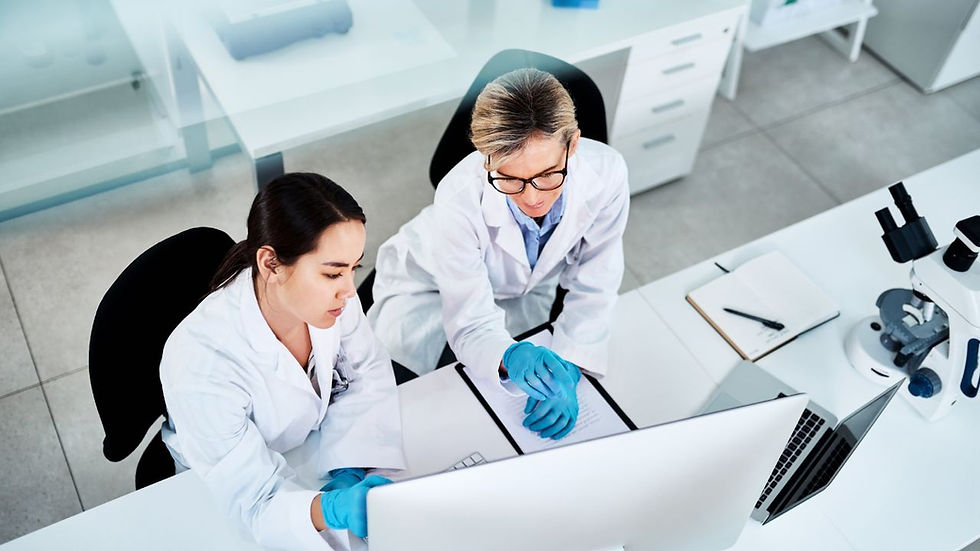 Two scientists in lab coats and gloves work at a computer in a bright lab, showing focus. Nearby are a microscope and open notebook.