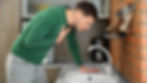 Man in green sweater leans over kitchen sink, holding chest in discomfort. Brick wall and kitchen appliances in background.