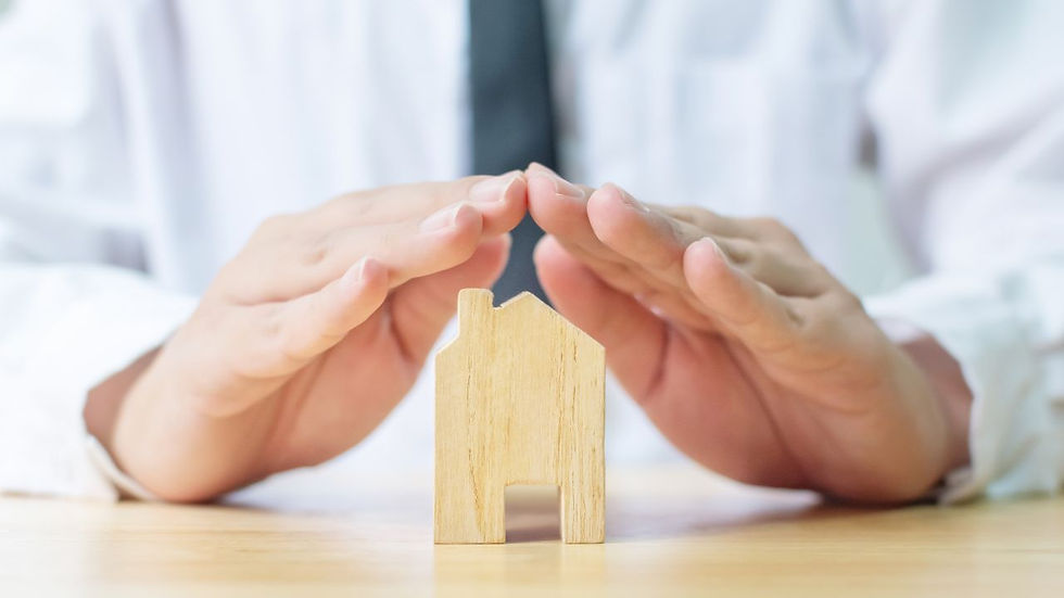Hands in a protective gesture over a small wooden house model on a table. Background shows a person in a white shirt and black tie.