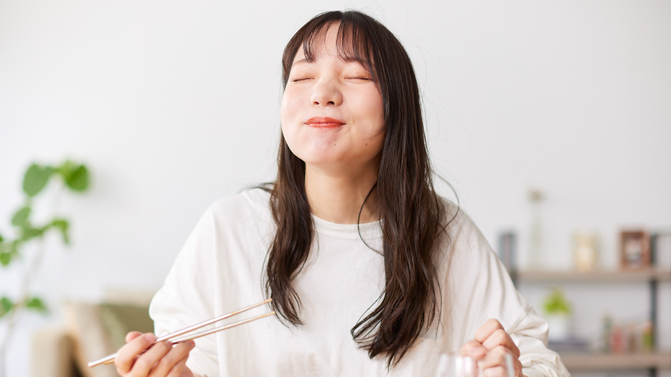 Smiling woman in a white shirt enjoys food with chopsticks in a bright room. Blurry plants and shelves in background.