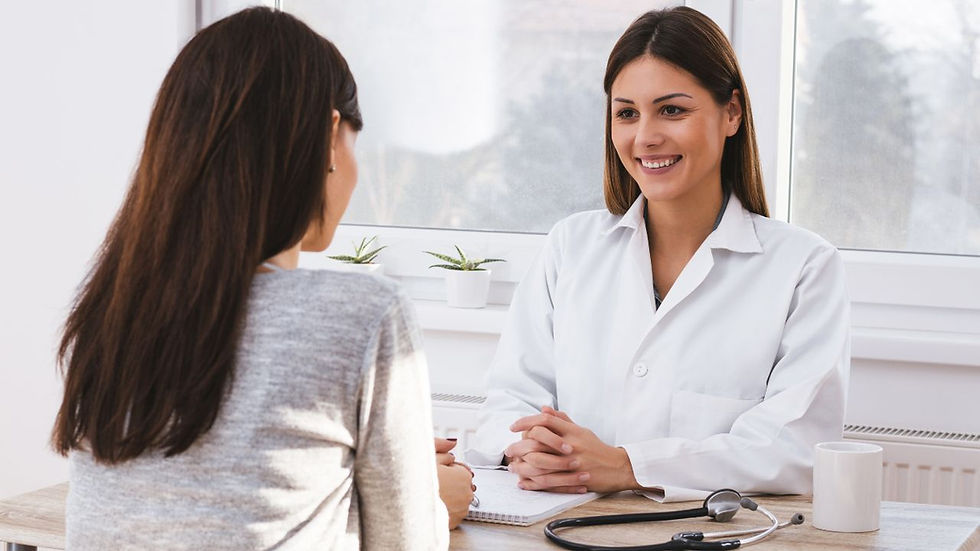 A doctor in a white coat smiles at a patient across a desk with a stethoscope and mug. Sunlight streams in through a window with plants.