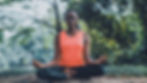 A woman in an orange tank top meditates in a serene forest setting, sitting cross-legged on a rock with eyes closed, exuding calmness.