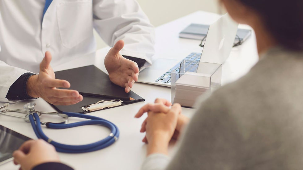 A doctor in a white coat talks to a patient across a desk. The doctor gestures with hands. A stethoscope, clipboard, laptop, and tissue box are on the desk. The patient is sitting with hands clasped.