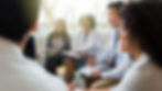 A diverse group of professionals sitting in a circle, engaged in a lively discussion. They are smiling and holding notebooks, with light streaming through a window in the background.