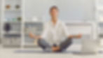 Person meditating on a mat, eyes closed, in a calm room with shelves and warm lights. Laptop nearby. Peaceful atmosphere.