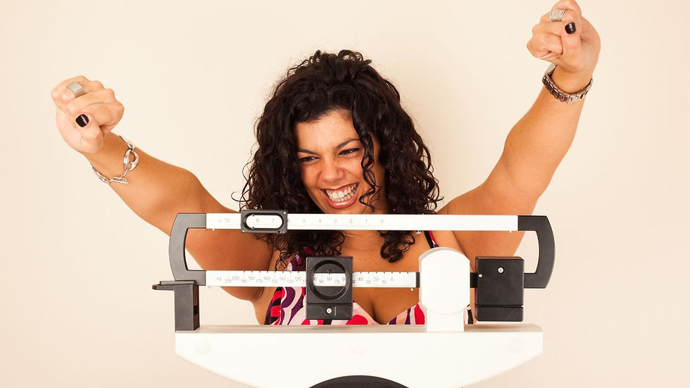 Woman joyfully raises fists in victory, standing behind a scale. She wears a patterned top, exuding happiness in a neutral background.
