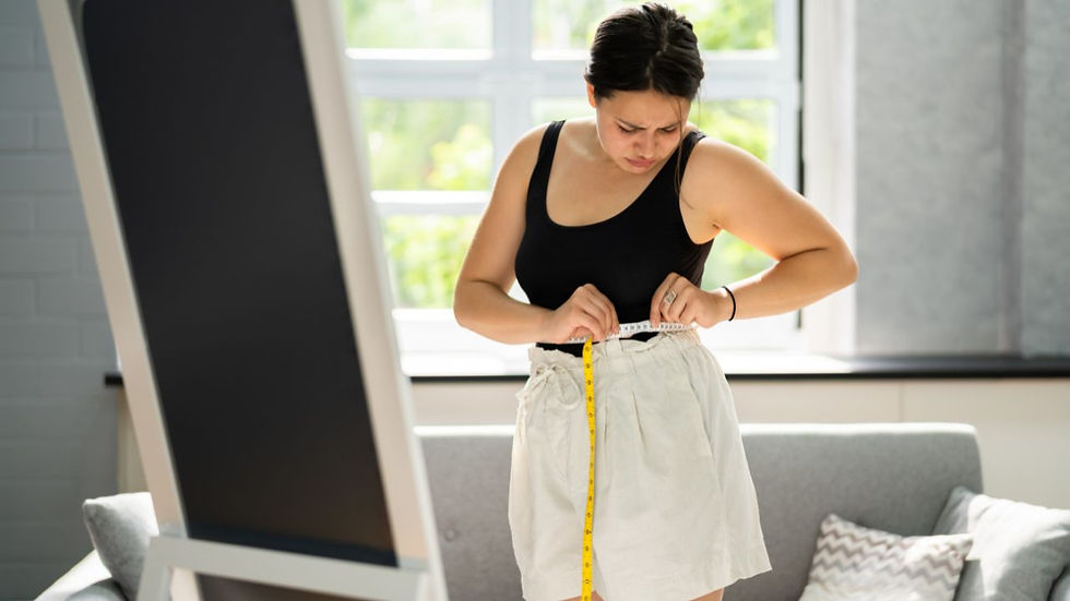Woman in black tank top measures her waist with a tape in a bright room with a couch. Concentrated mood, large window in the background.