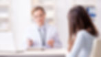 A doctor in a white coat consults a patient in an office. The doctor holds a stethoscope. Shelves with books are visible in the background.