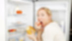 Woman eats tomato while holding bread in front of open fridge. Shelves display eggs, vegetables, and containers. Expression is surprised.