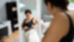 Woman in black top measuring her waist with a tape in front of a mirror. Room with sofa and shelf, natural light. Focused expression.