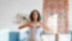 Woman practicing yoga in a room with a colorful patterned tapestry. She's focused, hands raised, wearing a white top, with plants on a nearby shelf.
