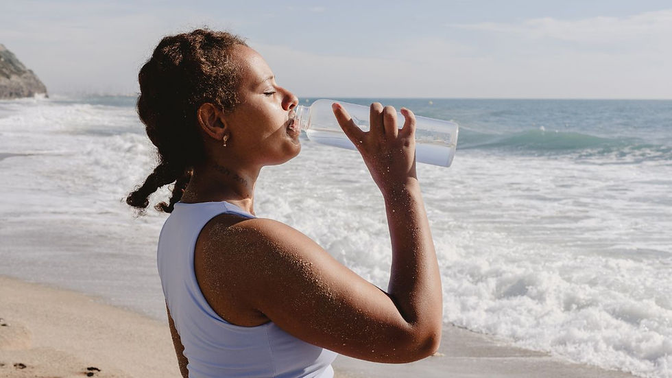 Woman in a blue tank top drinks from a bottle on a sunny beach. Waves in the background create a serene mood.