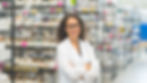 Smiling woman in a white lab coat stands with arms crossed in a warehouse with shelves of labeled boxes. Bright, organized setting.