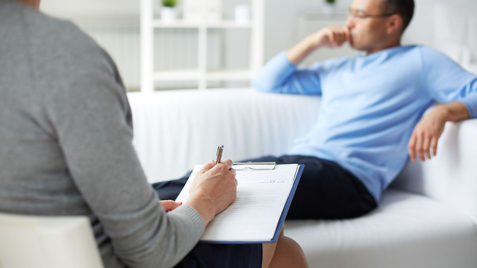 Therapist takes notes on a clipboard while a person in a blue sweater sits pensively on a white couch in a bright, calm room.