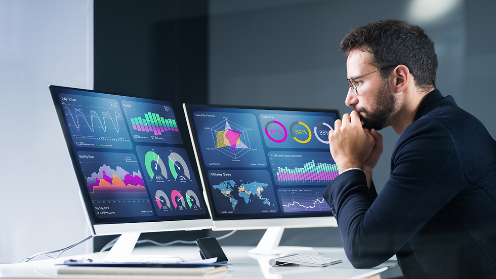 Man focusing on dual monitors displaying colorful data graphs and charts in a dark office setting, suggesting analysis and concentration.