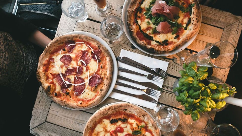Three pizzas with varied toppings on a wooden table with drinks, cutlery, and a basil plant. Cozy, inviting restaurant setting.
