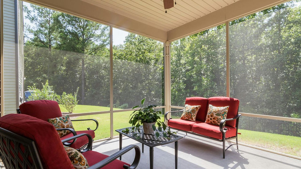 Screened porch with red cushioned furniture, floral pillows, and a table with a plant. Lush green trees and lawn visible outside.