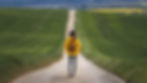 Person in yellow jacket and hat walks down a country road surrounded by green fields under a clear sky, creating a serene, adventurous mood.