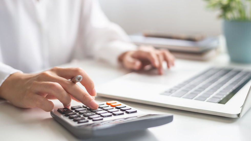 Hands using a calculator and laptop on a white desk. Background has a potted plant. Calm, focused workspace scene.