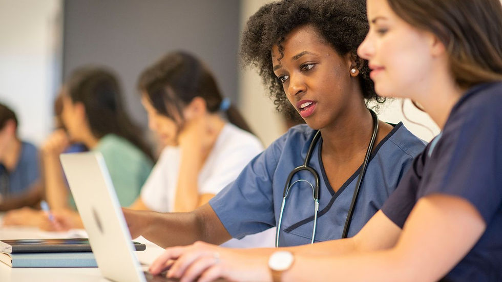Medical students collaborate on a laptop in a classroom, wearing scrubs. Focused and engaged, they sit at a bright table with books.